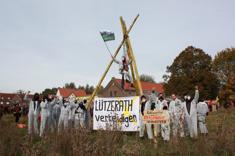 Menschen in weißen Anzügen stehen um einen Tripod herum und halten ein Transpi mit "Lützerath verteidigen"