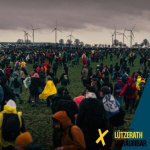 Viele Demonstrant*innen mit Regenklamotten und Rucksäcken auf einem Acker. Im Hintergrund Lützerath, PolizeiWannen und Windräder. Grauer, wolkenverhangener Wind