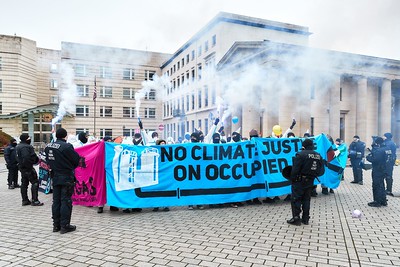 Ende Gelände Aktion vor einem Gebäude. Ein Block Demonstrierender ist umstellt von Cops. Die Menschen im Block sind größtenteils von Transpis verdeckt. Auf dem Fronttranspi steht in schwarz auf hellblauem Grund: No Climate Justice on Occupied Land